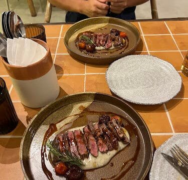 A person sits at a table in Siargao with two plates of sliced steak, sauce, and vegetables on a tiled surface.