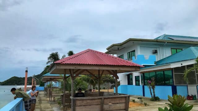 Wooden huts and benches by the sea in Siargao, near a blue and white building under a cloudy sky.