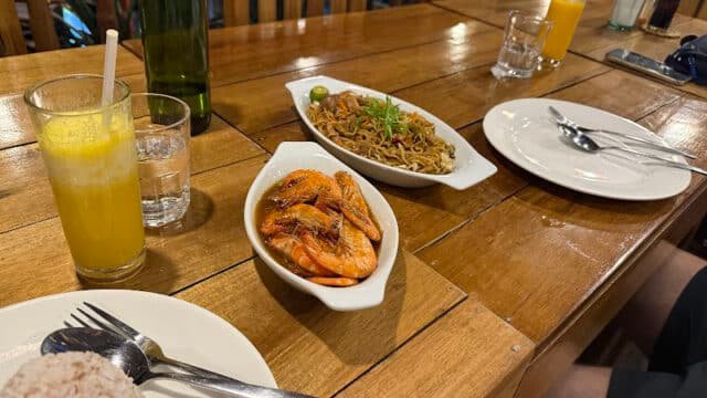 A wooden table in Siargao with plates of shrimp, noodles, rice, and drinks, all set for a delicious meal.