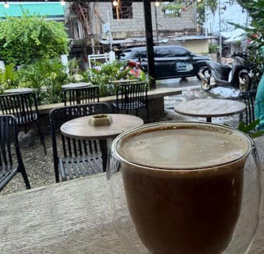 A glass mug of coffee on a table by a window, overlooking an outdoor café seating area in Siargao on a rainy day.