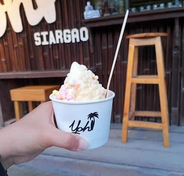 Hand holding a cup of ice cream with toppings in front of a wooden café, stool, and iconic Siargao sign.