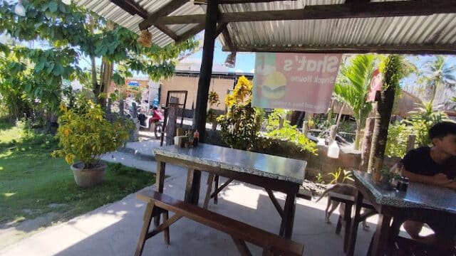 Outdoor café with metal tables and benches under a roof, leafy plants, and a sunny Siargao garden view.