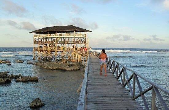 A woman walks on a wooden pier toward a stilted, open-air structure over the Siargao ocean at sunset.