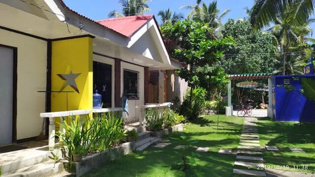Small tropical guesthouse in Siargao with yellow and white walls, lush garden, and palm trees under a sunny sky.