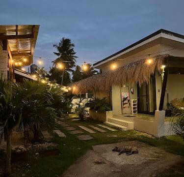 Cozy Siargao bungalow with string lights at dusk, palm trees swaying, and a dog lying outside.