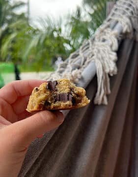 A hand holds a half-eaten chocolate chip cookie in front of a hammock and palm trees outdoors in sunny Siargao.