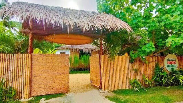 Entrance to a bamboo-fenced Siargao hostel with a thatched roof, lush tropical plants, and a sign reading Siargao Tropic Hostel.