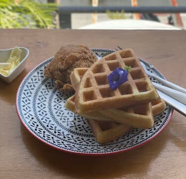 Plate of waffles topped with a purple flower, served with fried chicken and butter on a rustic Siargao wooden table.