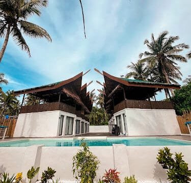 Modern Siargao tropical villas with pointed roofs by a swimming pool, surrounded by palm trees and lush greenery.