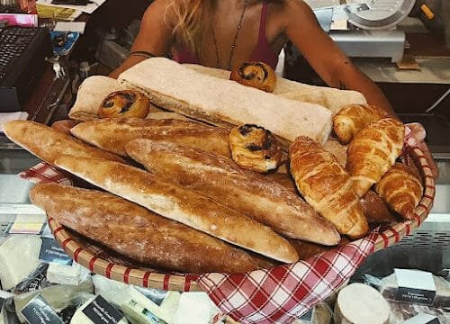 A woman in Siargao holds a large basket of baguettes, croissants, and pastries over a deli counter with assorted cheeses.