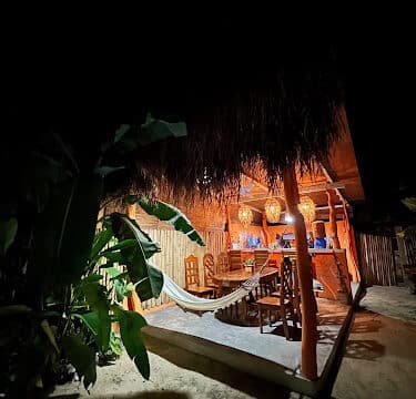 A cozy, thatched outdoor bar in Siargao with chairs and a hammock, warmly lit at night, surrounded by lush tropical plants.
