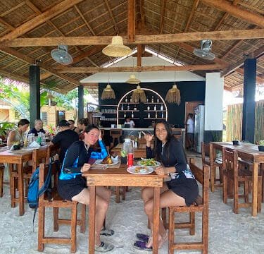 Two women smile while dining at a Siargao beachside restaurant with sandy floors and wooden furniture.