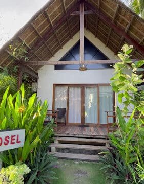 A small thatched-roof cottage with a porch, surrounded by green plants in Siargao, with a sign partially reading Hostel.