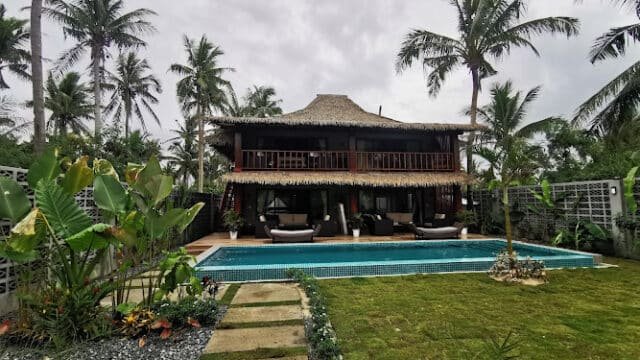 Two-story tropical Siargao villa with thatched roof, pool, palm trees, and lush green garden under a cloudy sky.