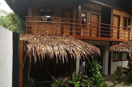 Two-story tropical Siargao house with thatched roof, wooden balcony, and lush green plants in front.