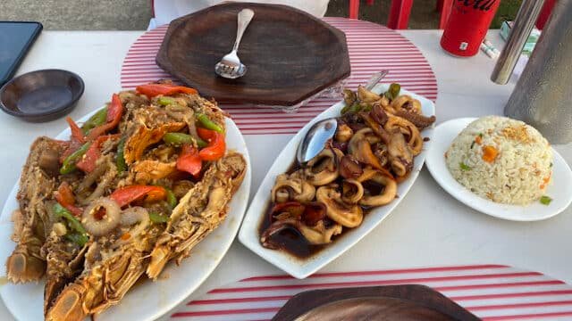 Seafood dishes, fried rice, and a can of Coke on a white table with red striped placemats outdoors in sunny Siargao.