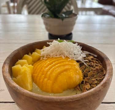 A wooden bowl with sliced mango, pineapple, granola, and shredded coconut on a light table; Siargao vibes with a plant in the background.