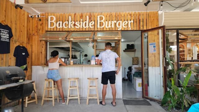 Two people stand at the counter of Siargao’s wooden burger stand, Backside Burger, with stools out front.