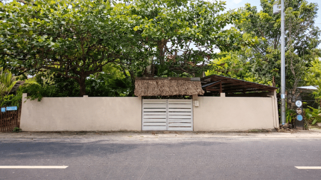 A white gate and wall with trees and a thatched roof above, seen from across a Siargao road on a sunny day.