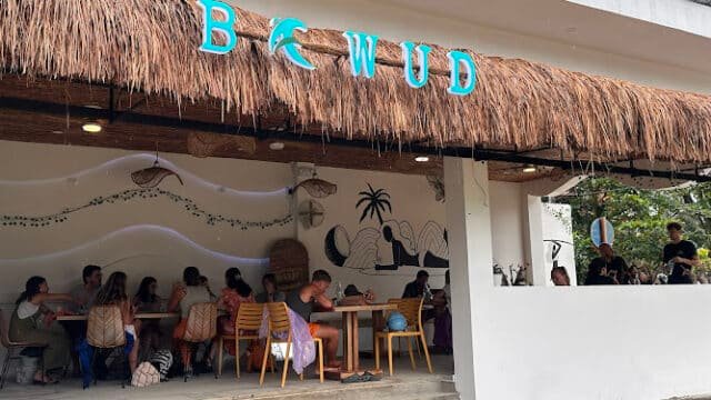 People sitting and dining under a thatched roof at a Siargao cafe with a sign that reads BCWUD.