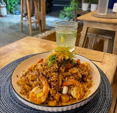 A bowl of seafood pad Thai with shrimp and vegetables, served Siargao-style on a table with a glass of sparkling water.