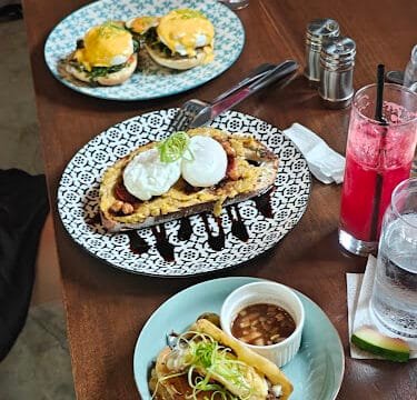 Three plates of brunch food with drinks on a wooden table in Siargao, featuring eggs benedict, waffles, and tacos.