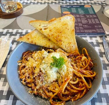 A bowl of spaghetti with tomato sauce, grated cheese, and parsley, served Siargao-style with two slices of garlic bread.