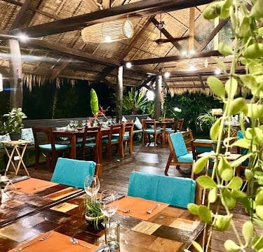 Open-air Siargao restaurant with wooden tables, turquoise chairs, lush plants, and warm lighting under a thatched roof.