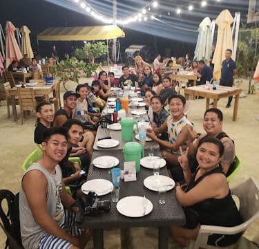 A group of people smiling and gathered around a long, outdoor dinner table in Siargao at night with string lights above.