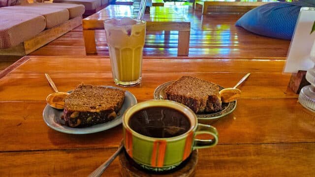 Two plates with banana bread, a cup of coffee, and an iced latte on a wooden table in a cozy Siargao café setting.