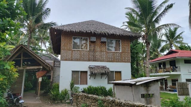 A two-story tropical house with bamboo walls, thatched roof, and Siargao palm trees in the background.