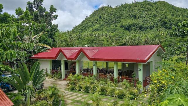A building with a red roof in Siargao, surrounded by lush greenery and hills under a partly cloudy sky.