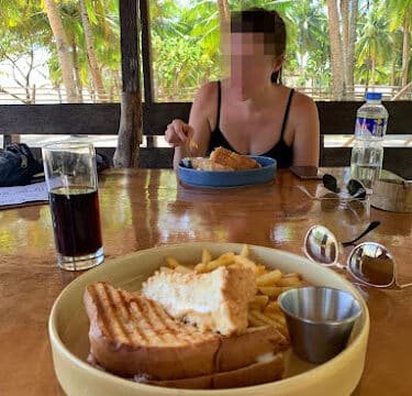 A person at a beachside table in Siargao enjoys a sandwich, fries, and a drink, with palm trees swaying in the background.