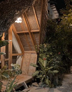 A cozy, wooden A-frame hut in Siargao at night, surrounded by plants and illuminated by a warm hanging light.