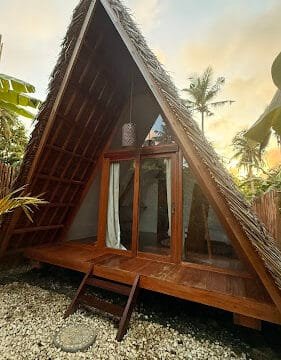A-frame wooden cabin with glass doors and thatched roof, surrounded by plants and a stone path in scenic Siargao.