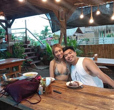 A smiling couple sits at a rustic Siargao outdoor restaurant table with food and drinks in front of them.