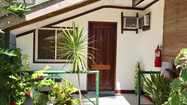 Small house entrance in Siargao with wooden door, lush greenery, potted plants, and a window air conditioner on a sunny day.