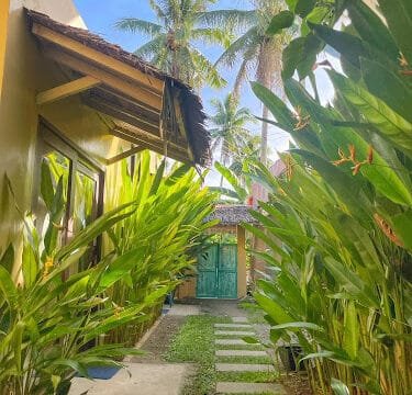 Stone path between tropical plants and yellow buildings in Siargao, leading to a teal gate under palm trees and blue sky.