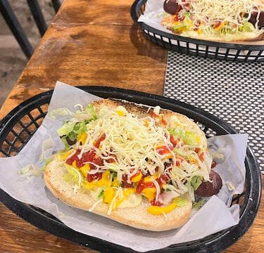 A hot dog topped with shredded cheese, lettuce, ketchup, and mustard on a wooden table in sunny Siargao.