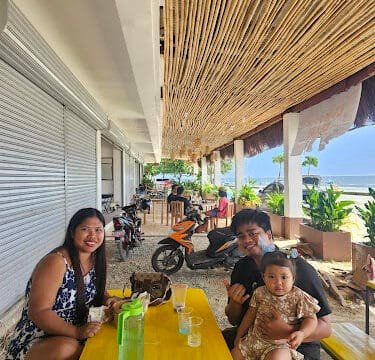 A smiling family sits at a yellow table under a bamboo roof at an outdoor Siargao cafe with parked motorcycles nearby.