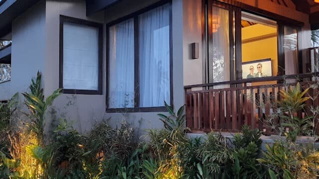 Modern Siargao house with large windows, wooden balcony, and garden plants beautifully lit by outdoor lights at dusk.