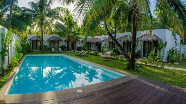 Sunny poolside area with palm trees, loungers, and thatched-roof cabanas in a lush Siargao tropical garden setting.