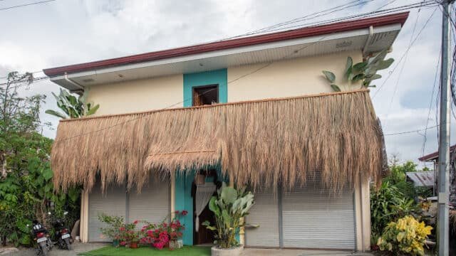 Two-story Siargao house with thatched roof awning, lush plants, and closed metal shutters on a cloudy day.