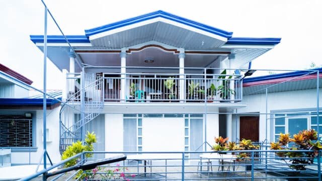 Two-story Siargao house with a balcony, outdoor plants, metal railings, and blue accents on the roof and walls.