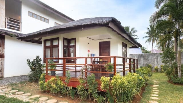Small modern bungalow with a wooden porch, nestled among plants and a stone path in a lush Siargao tropical garden.