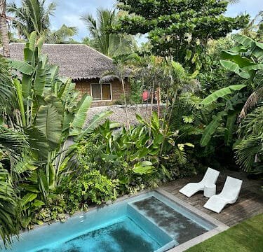 A tropical Siargao backyard with a pool, lush greenery, two white lounge chairs, and a thatched-roof house.