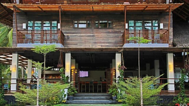 Two-story wooden building in Siargao with a balcony, open seating area below, and lush plants in front.