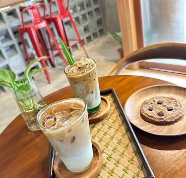 Two iced lattes and a chocolate chip cookie on a wooden table in a cozy Siargao café with red stools in the background.
