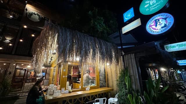 Outdoor night view of a small, tiki-style Siargao bar with patrons and bright neon restaurant signs above.