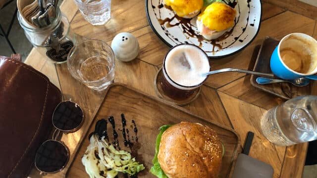 Wooden table in Siargao with a burger, eggs benedict, drinks, sunglasses, and utensils arranged on top.
